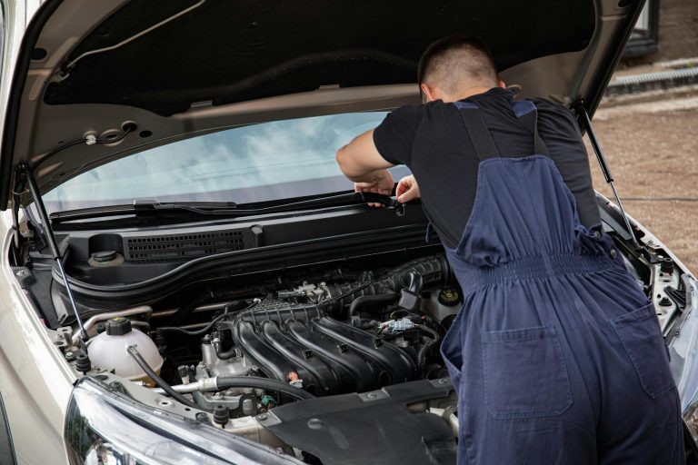 A mechanic in blue overalls working on a car engine as part of maintenance routine.
